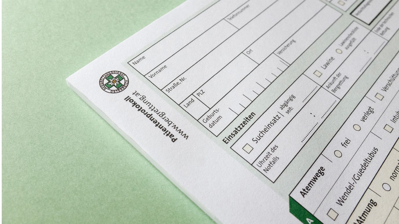 Close-up of an emergency documentation form. On a green background. The form includes fields for name, address, birth date, and checkboxes for Einsatzzeiten (operation times) and Atemwege (airways). The logo of the Tyrolean Mountain Rescue Service is visible in the top left corner.