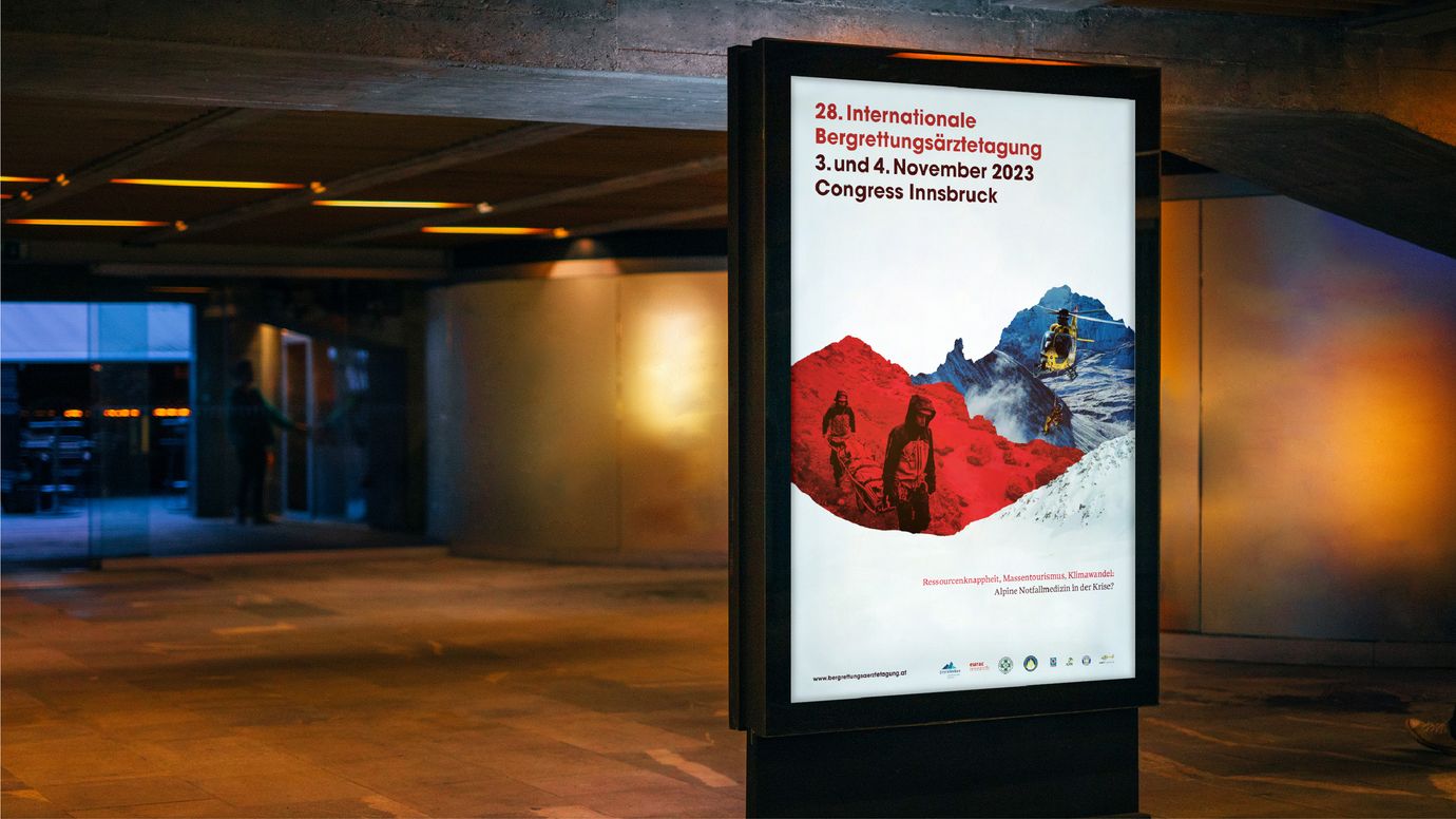 Backlit City Light poster in a pedestrian underpass. The poster shows the title “International Mountain Rescue Doctors Conference and November Congress Innsbruck.” A silhouette-like collage in the form of mountain ranges, which included the famous “Frau Hitt” above Innsbruck, shows two images: on the left, in red: two mountain rescuers carry an alpine rescue stretcher down a hiking trail; on the right: a yellow helicopter with a rescuer on a rope flies between mountains.